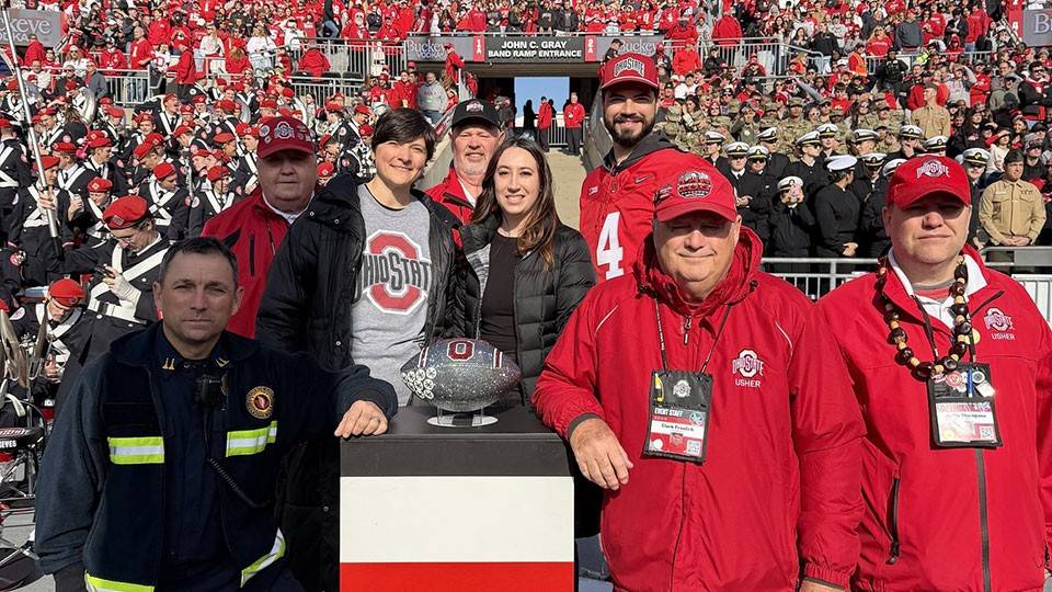 Ohio state football staff, paramedic, and Red Cross volunteers stand next to the Ohio state football trophy in the Ohio state football stadium that's filled with fans.