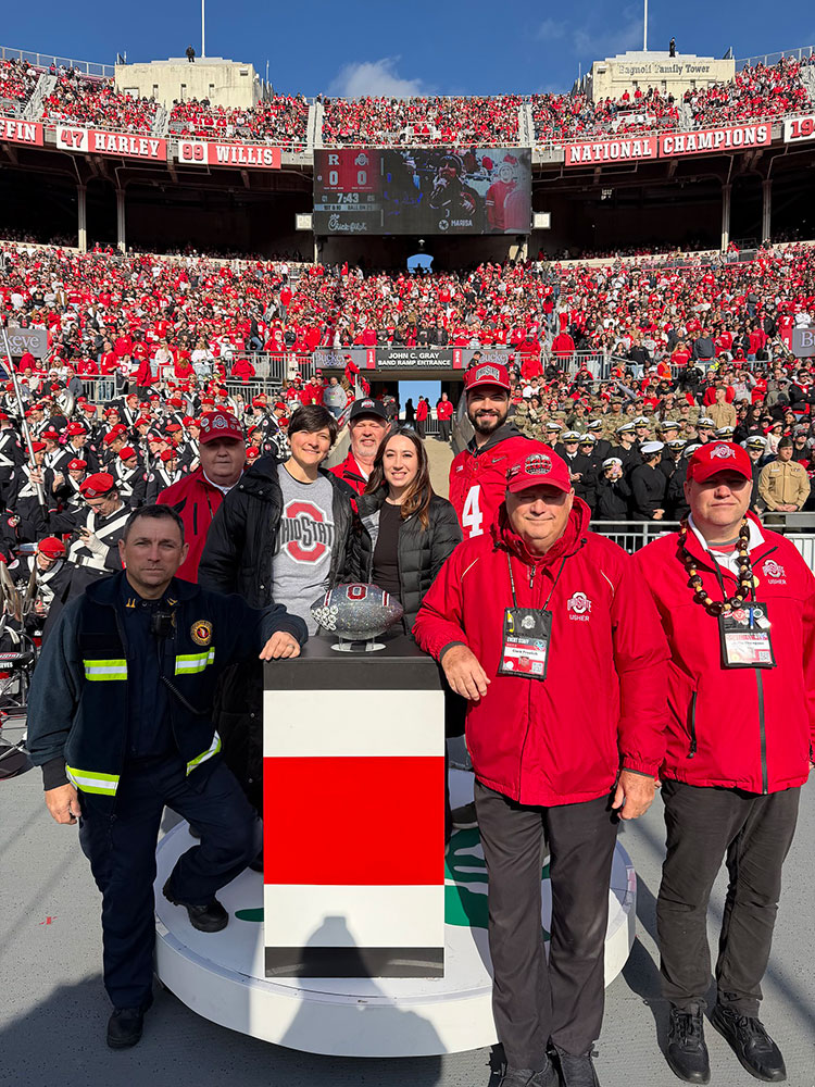 Ohio state football staff, paramedic, and Red Cross volunteers stand next to the Ohio state football trophy in the Ohio state football stadium that's filled with fans.