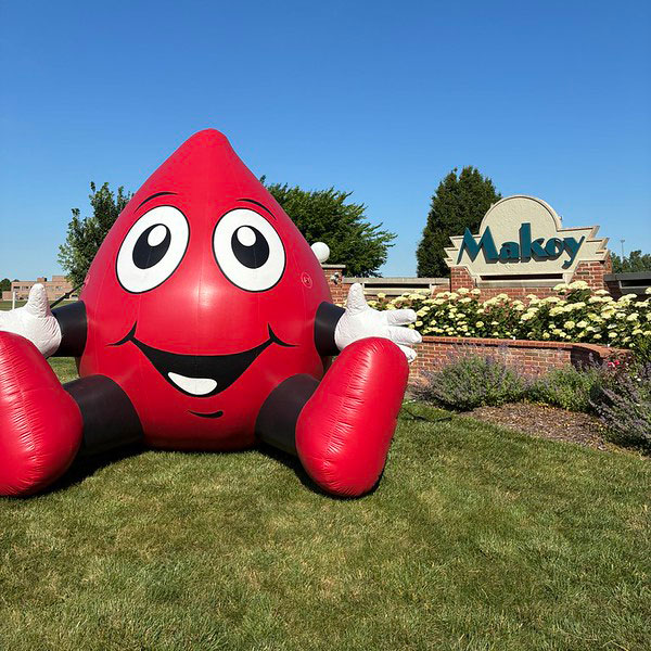 Rodney the red cell mascot, that's shaped like a blood drop, sitting on grass in front of a Makoy sign.