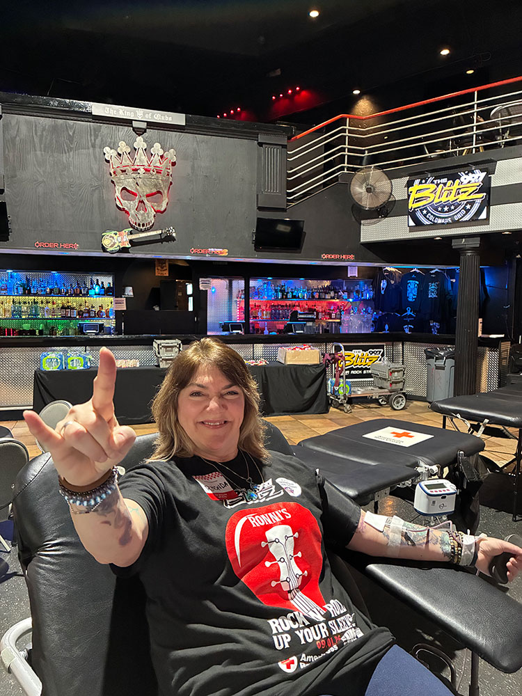 a woman sitting in a chair donating blood while smiling and giving the sign of the horns at the King of Clubs concert venue.