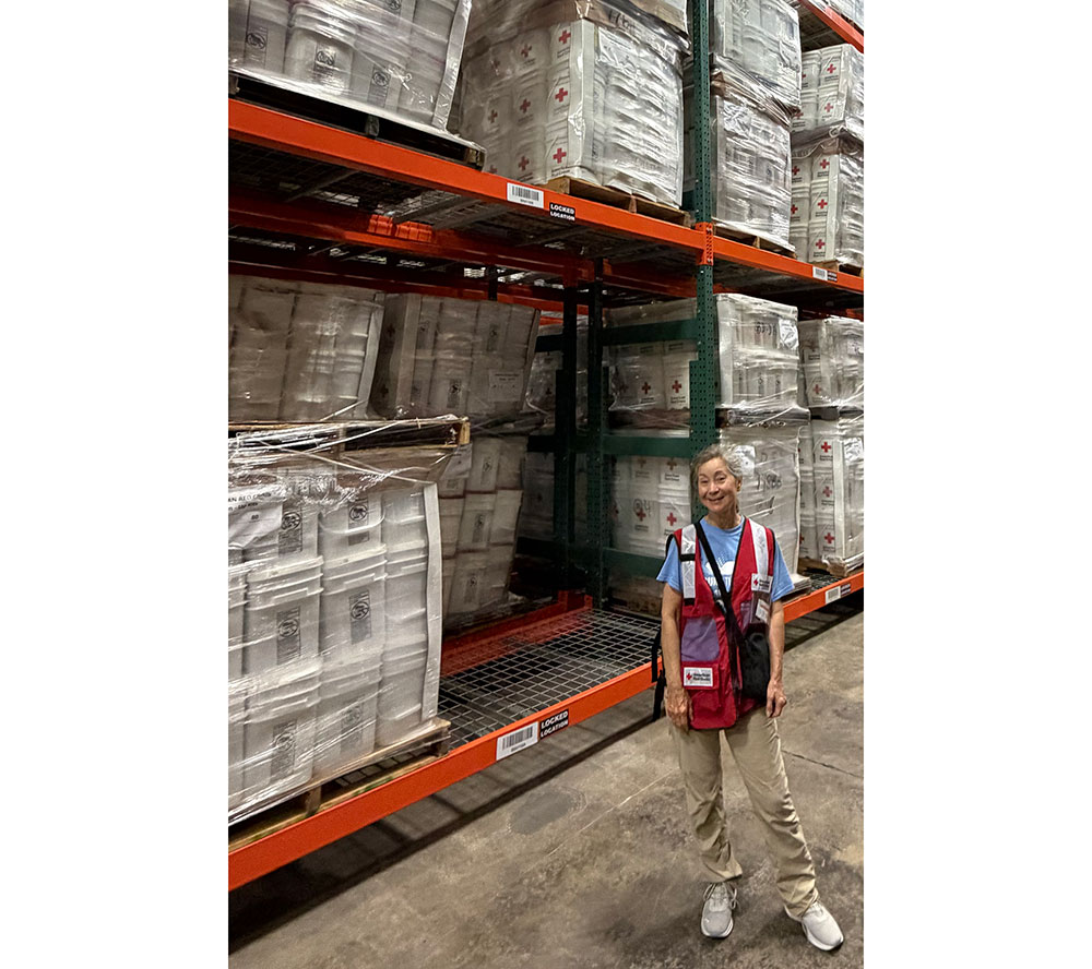 Pam Benton standing in a Red Cross warehouse with pallets filled with Red Cross boxes.