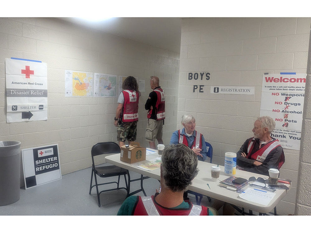 People sit at a table and stand in a hallway while waiting for people affected by a wildfire. Several signs indicate where to go to find a Red Cross shelter.