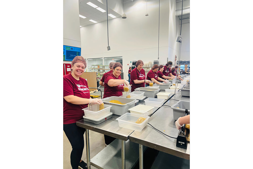 Workers in maroon shirts and hair nets pack dry pasta into containers on a stainless steel assembly line.