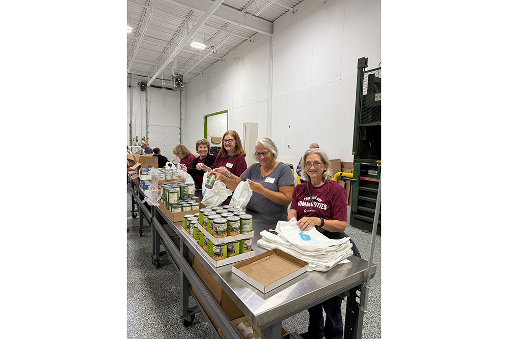 A group of people in a warehouse smiling while packing canned goods and supplies into bags on a long table.