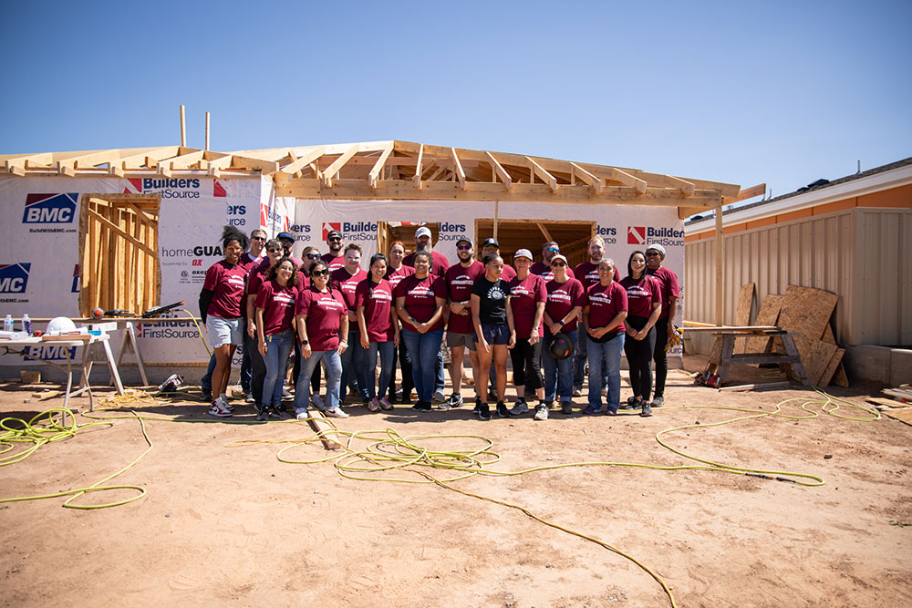 A group of people in matching maroon shirts stand in front of a partially constructed house under a clear blue sky.