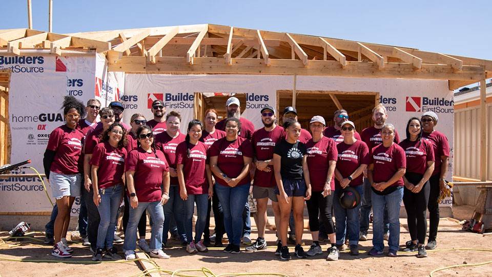 A group of people in matching maroon shirts stand in front of a partially constructed house under a clear blue sky.