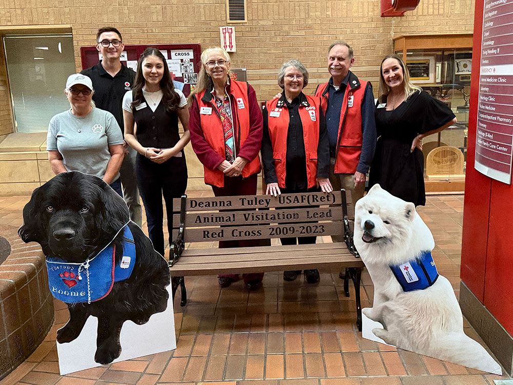 Red Cross staff members standing next to bench that's dedicated to Deanna Tuley, and two cut out pics of service dogs.