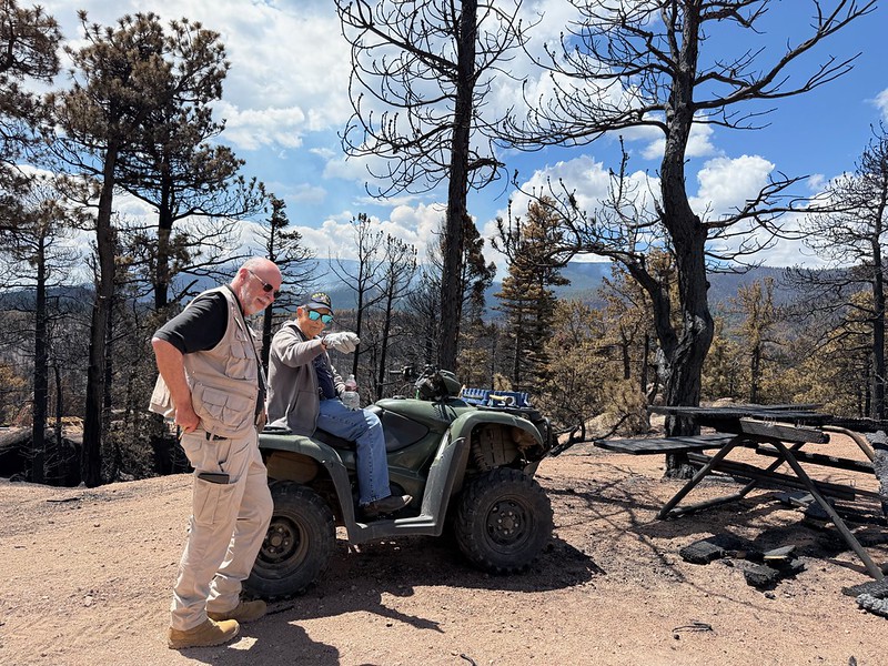 A man on an off-road vehicle points at something, while another man in a Red Cross vest and sunglasses looks at what is being pointed at. Behind them are burnt trees and a burnt picnic table.