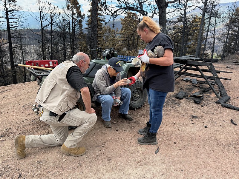 A woman hands a man sitting down an item recovered from the remains of a burnt-out house. A second man in a Red Cross vest kneels next to the man sitting down.