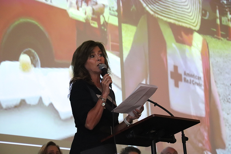 A woman with a microphone in hand speaking on stage, with a collage of Red Cross photos behind her