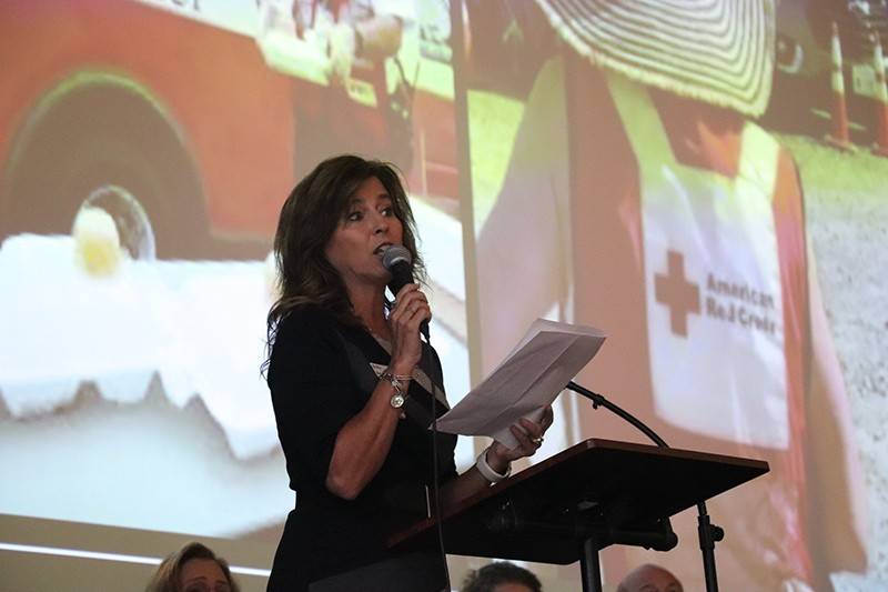 Woman with a microphone in hand speaking, with a collage of Red Cross photos behind her