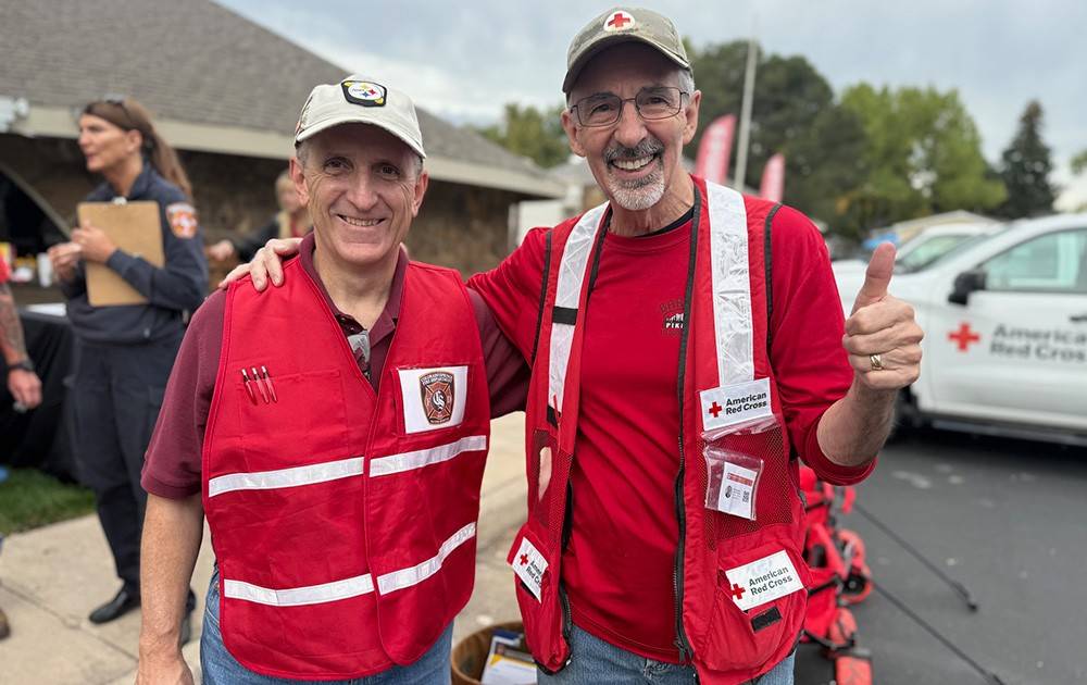 Two men in Red Cross vests stand together, with one giving a thumbs up while smiling