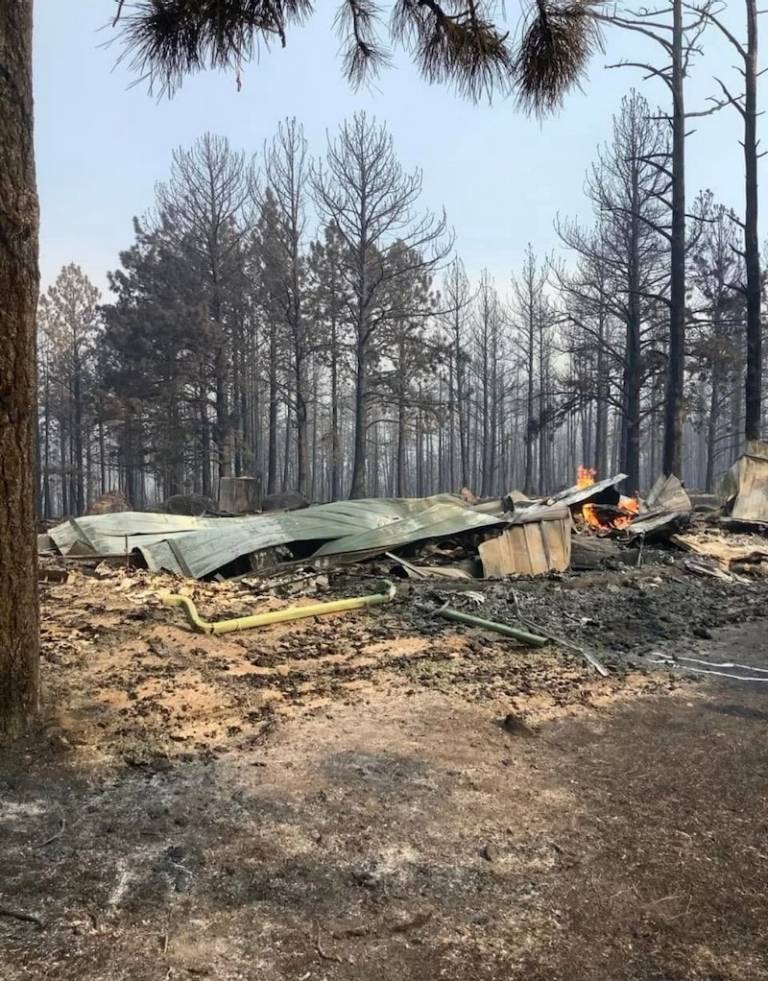 Remains of a home that burned down with only steel roof left, flames in background