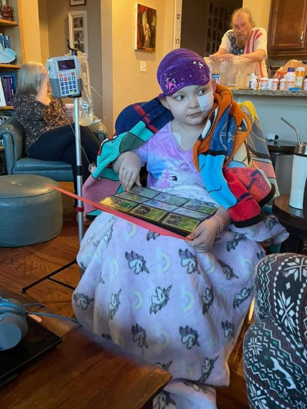 A girl sits in a living room, holding a binder of collectible cards, while she talks to someone out of frame. An IV bag and tube are next to her, with a bandage on her face and a tower wrapped around her shoulders.