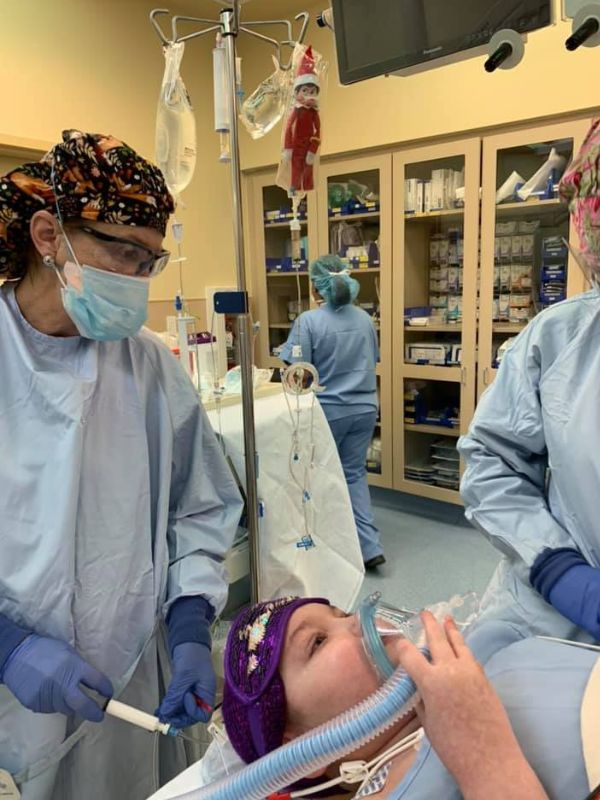 A young girl, with a breathing mask covering her nose and mouth, is surrounded by several people dressed in medical equipment and face masks. She is lying down on a table while receiving medical treatment..