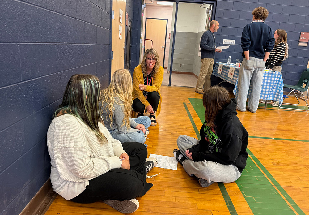 A woman in a yellow sweater talks with three students sitting on the floor