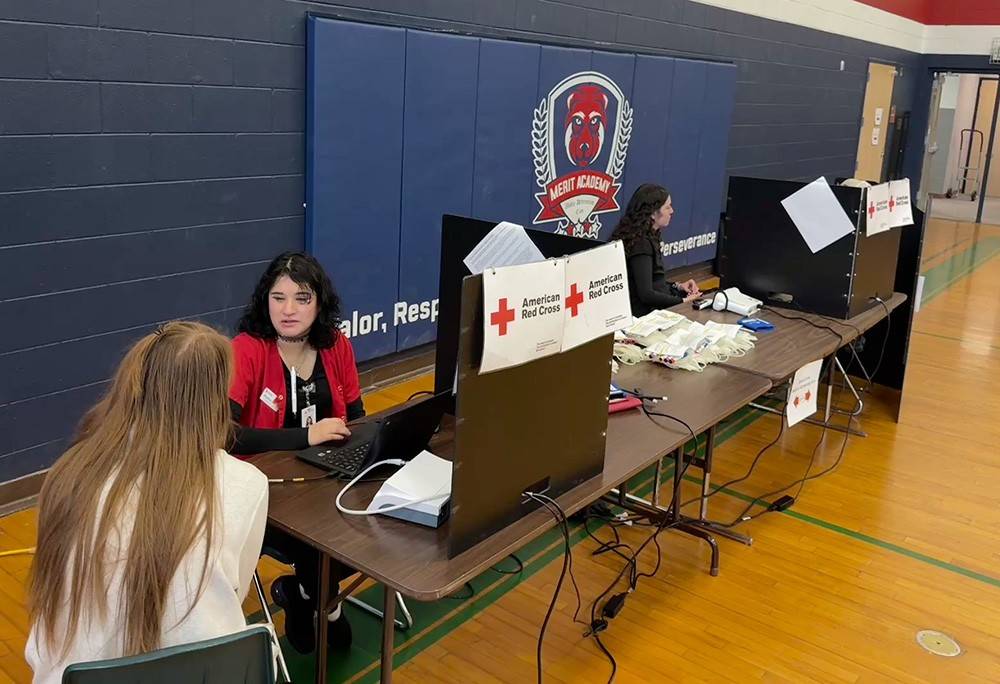 Red Cross phletbotomists conduct pre-donation screenings at Merit Academy. The Merit Academy logo is visible in the background.