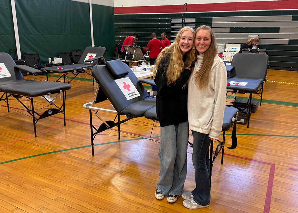 A woman and her daughter standing in front of multiple Red Cross blood donation tables inside of a gym at a blood drive.