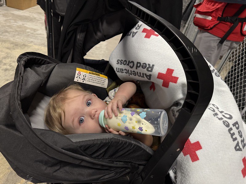 Baby drinks from a bottle while wrapped in a Red Cross blanket