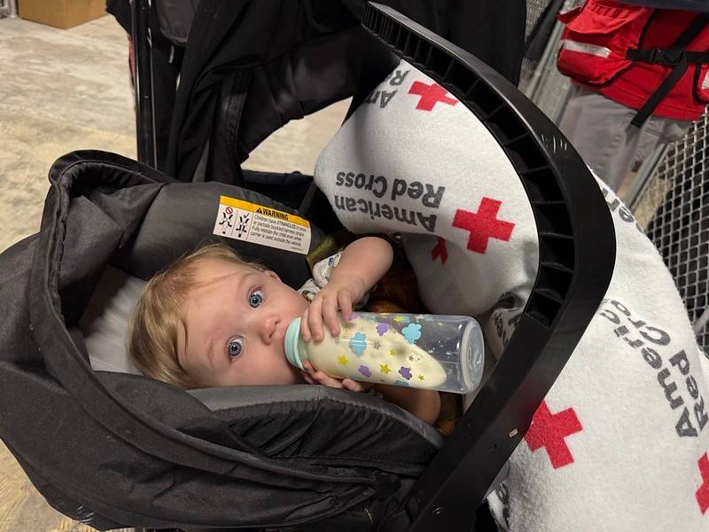 Baby drinks from a bottle while wrapped in a Red Cross blanket
