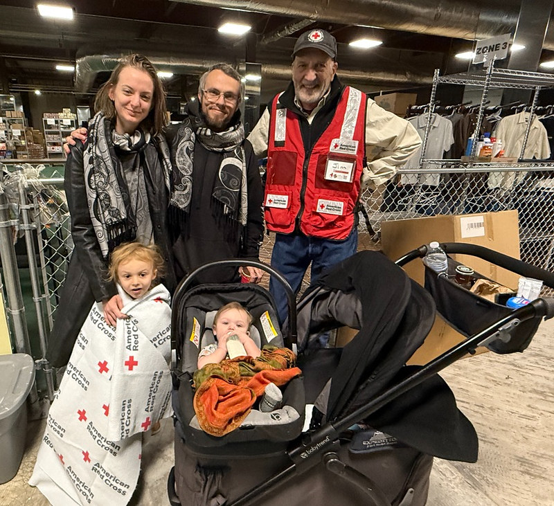 Family stands with a Red Cross volunteer in a shelter. A stroller is at their side