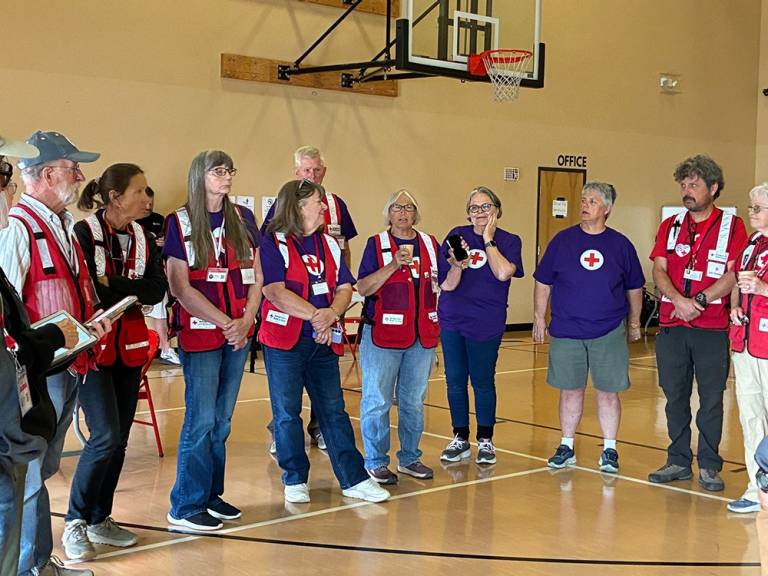 Red Cross volunteers, including some with purple shirts and some in red vests, stand in a group inside of a gym as one person speaks.