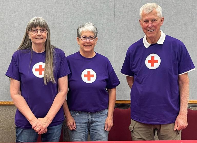 Three volunteers - two women and one man - stand together behind a Red Cross-branded table. All three are wearing purple shirts.