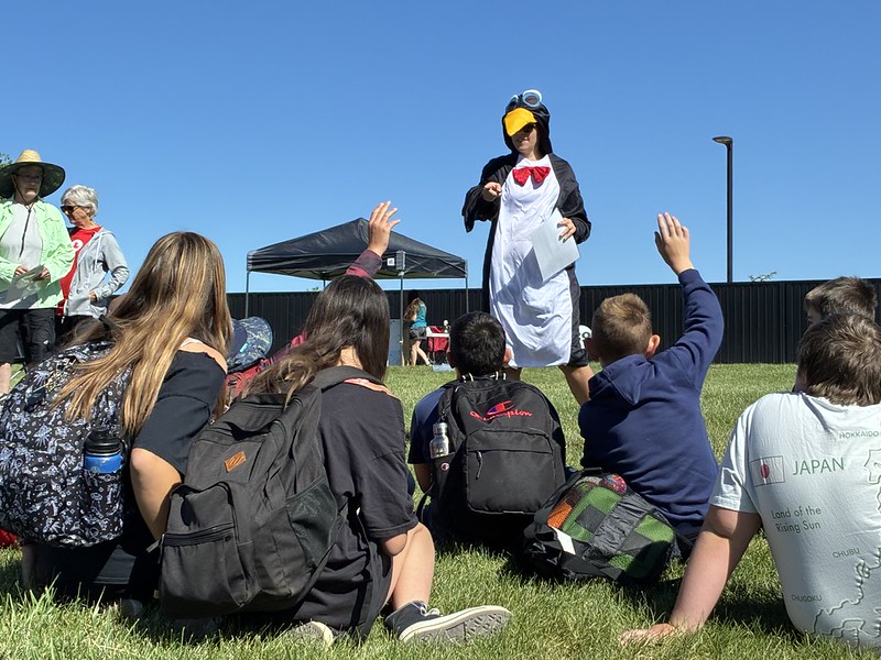 Pedro the Penguin gives a presentation outdoors in front of several students sitting on the ground