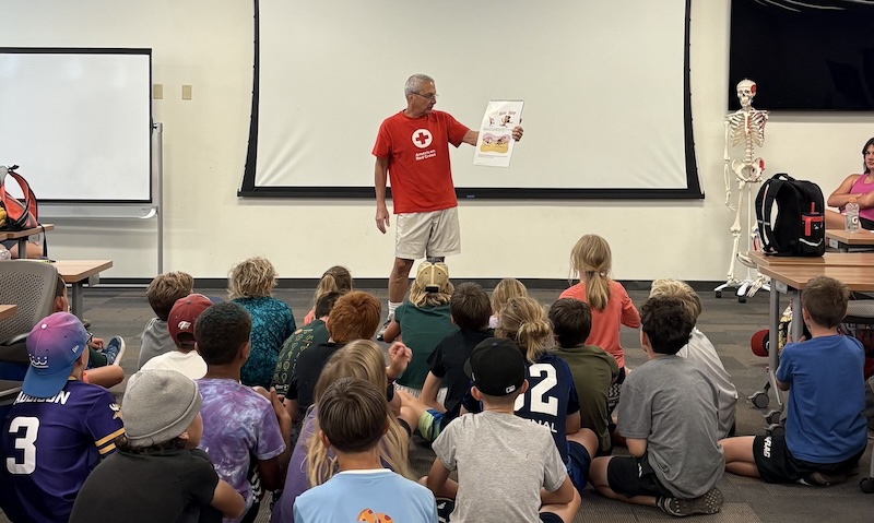 A Red Cross volunteers gives a presentation in a room in front of several students sitting on the ground