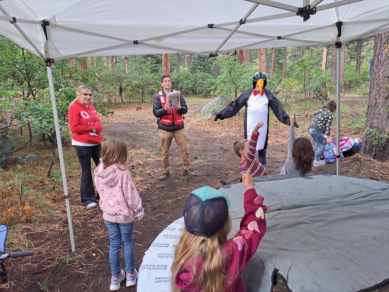 Red Cross volunteer talking to students alongside Pedro in an outdoor setting. Some students are raising their hands in response to a question.