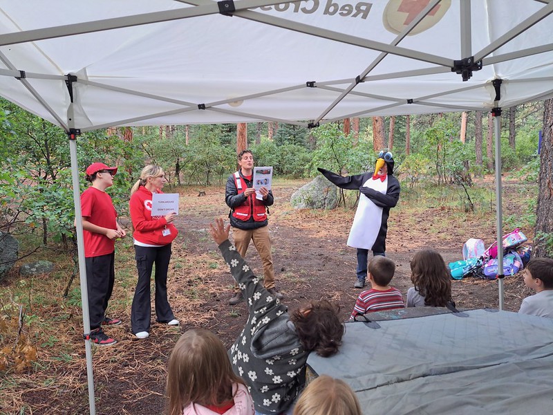Red Cross volunteer talking to students alongside Pedro in an outdoor setting. Some students are raising their hands in response to a question.