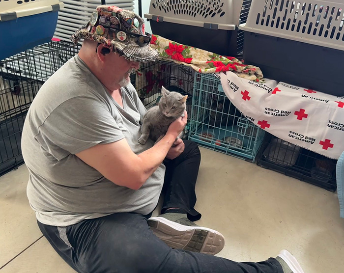 A man sitting down, holding a gray cat in his arms; a Red Cross blanket is visible on top of the crates