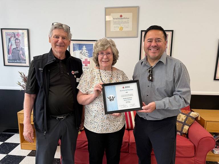 Two men stand next to a woman, holding a certificate of appreciation. All are smiling. Red Cross memorabilia is in the background.