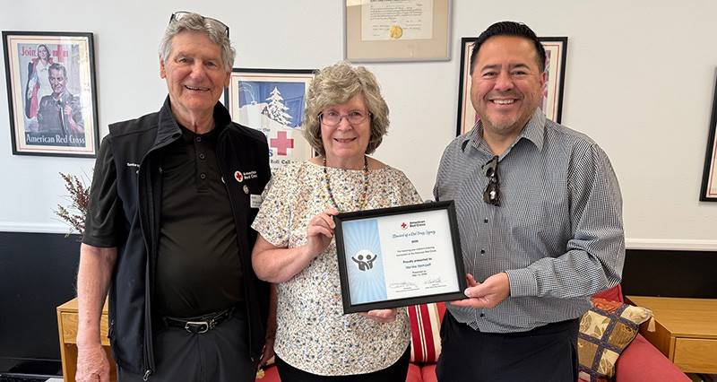 Two men stand next to a woman, holding a certificate of appreciation. All are smiling. Red Cross memorabilia is in the background.