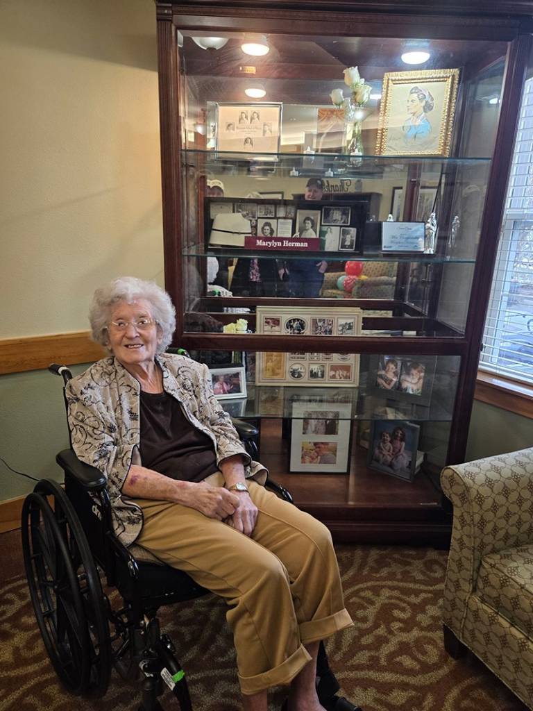 A woman in a wheelchair sits next to a large display of memorabilia and mementos honoring her nursing service