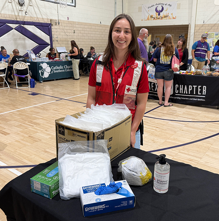 A Red Cross volunteer standing behind a table with personal protective equipment like masks and gloves in front of her.