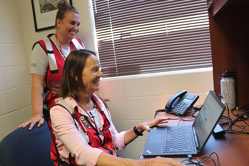 Woman sitting at a desk in front of a computer with another woman in a Red Cross vest behind her, smiling