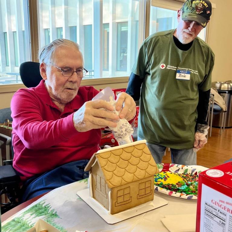 A man frosts a gingerbread house while another man observes.