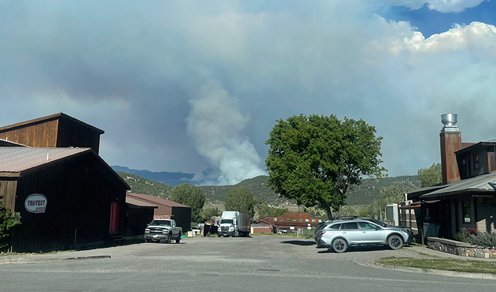 Smoke rises from a fire burning in the distance beyond mountains, with cars and buildings in the foreground