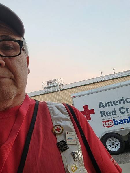 Man dressed in Red Cross vest and gear stands outside a building with a Red Cross trailer behind him