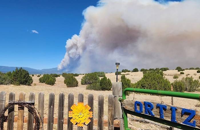 Smoke rises in the distance behind trees and mountains. In the foreground is a fence along with letters spelling out "Ortiz" on the fence