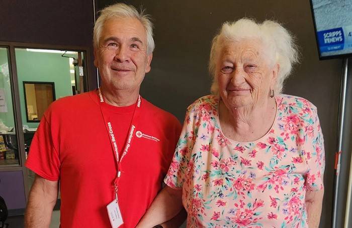 A Red Cross volunteer in a red shirt stands next to an older woman in a pink shirt