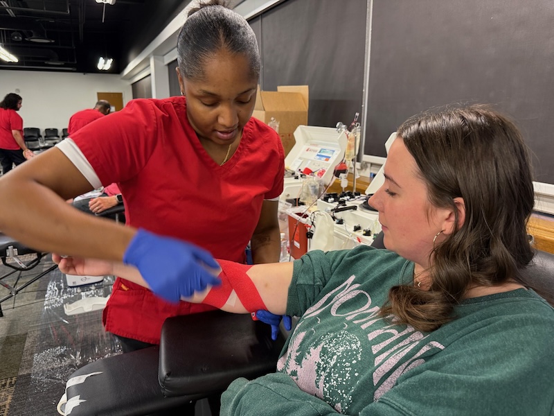 A woman donates blood, with a Red Cross phlebotomist standing next to her