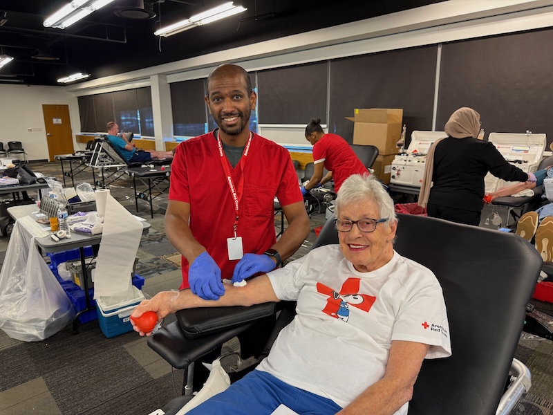 A woman donates blood, with a Red Cross phlebotomist standing next to her