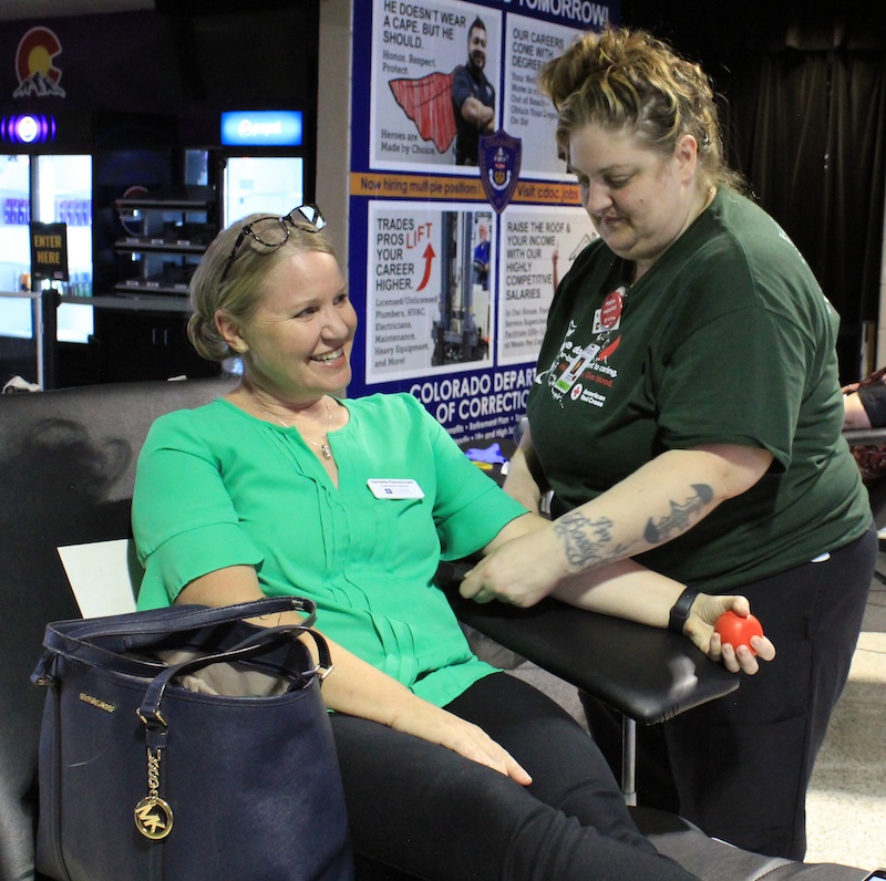 A woman in a green shirt prepares to donate blood with a smile on her face, with a Red Cross phlebotomist standing next to her