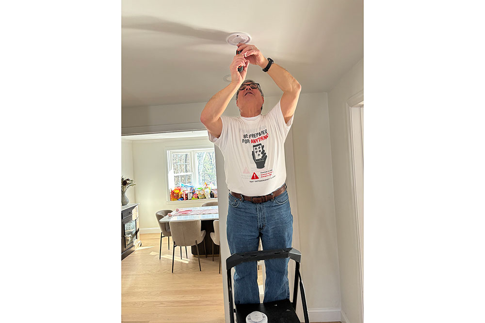 Larry Berman in jeans and a t-shirt stands on a step ladder, installing a smoke alarm on the ceiling. The room is well-lit with a dining table visible in the background.