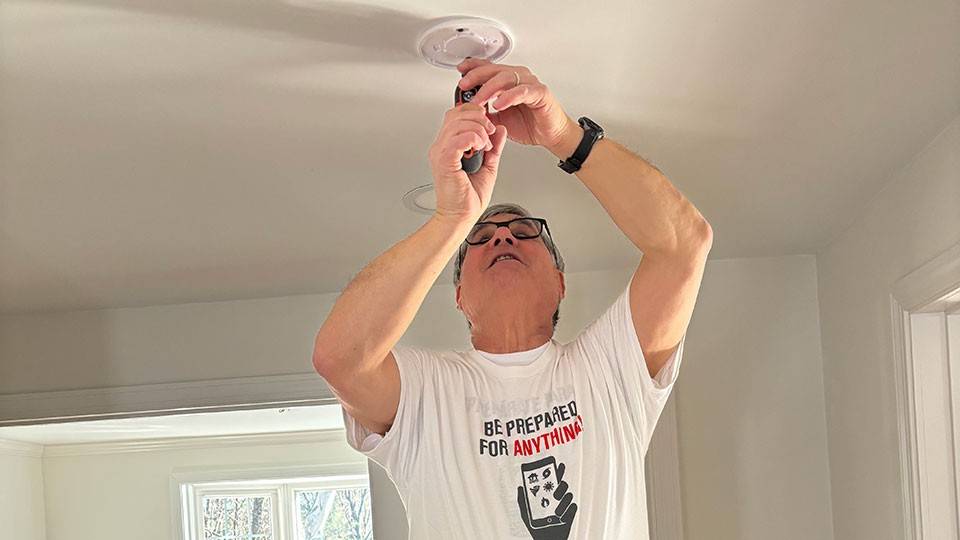 Larry Berman in jeans and a t-shirt stands on a step ladder, installing a smoke alarm on the ceiling. The room is well-lit with a dining table visible in the background.