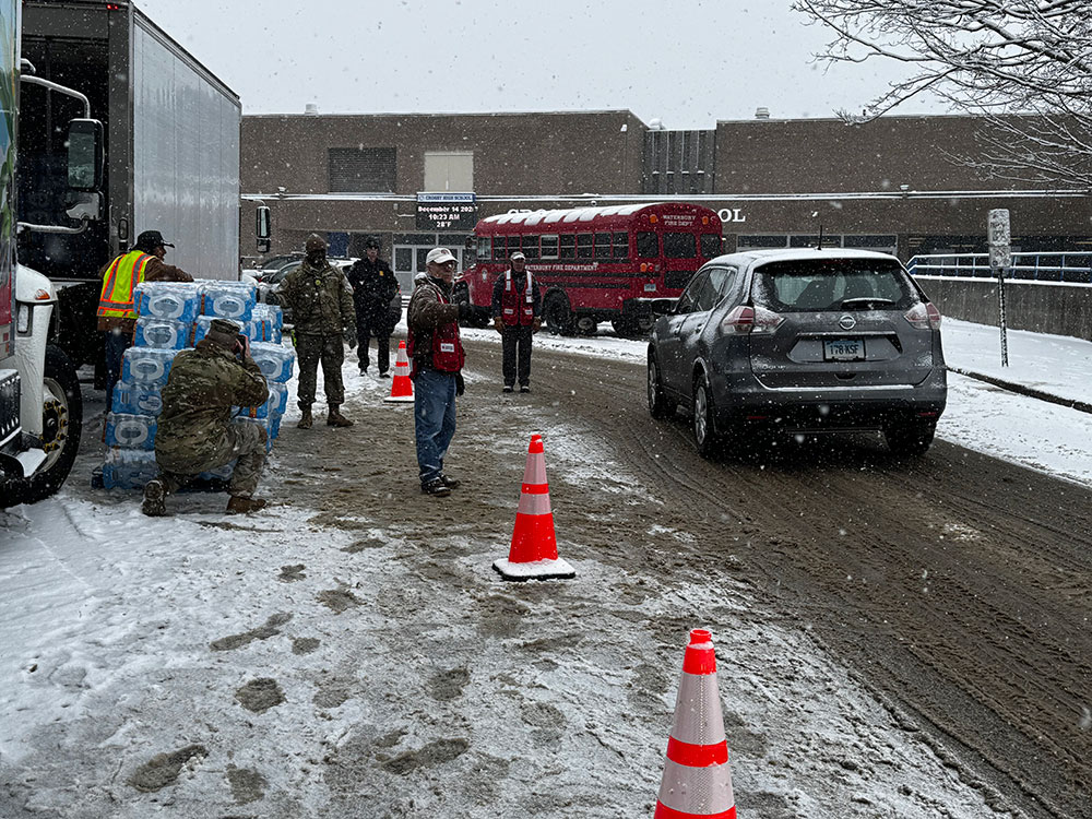 Red Cross volunteers directing traffic in the snow with a moving truck and a stack of bottled waters on the side of the road.