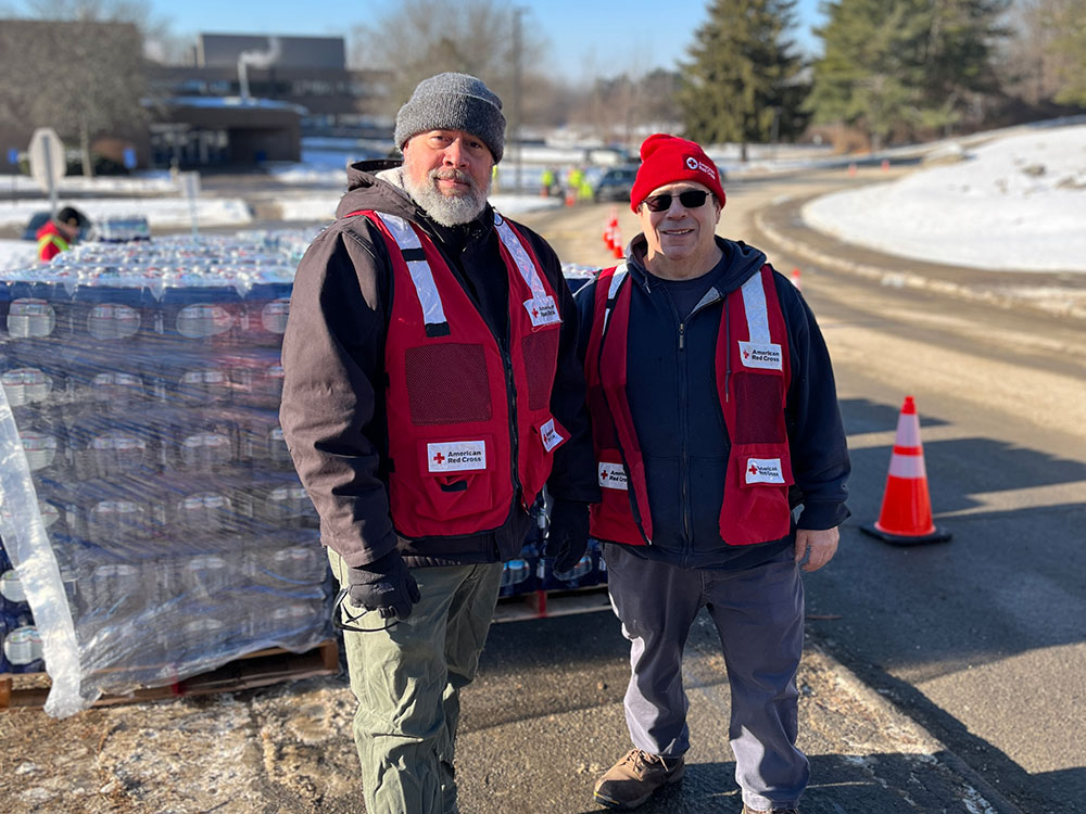 TTwo Red Cross volunteers standing next to a stack of bottled waters on the side of the road.
