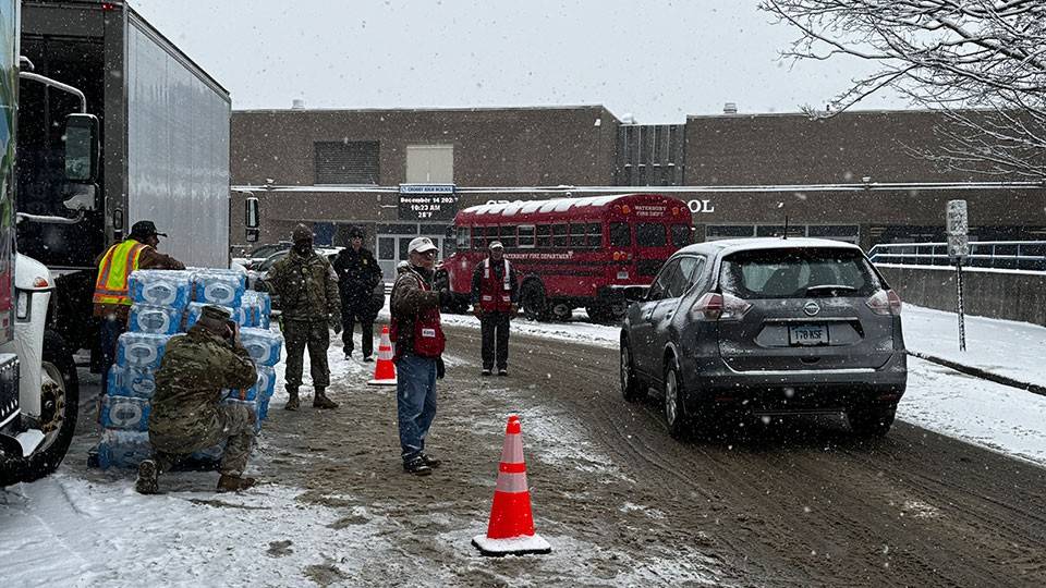Red Cross volunteers directing traffic in the snow with a moving truck and a stack of bottled waters on the side of the road.